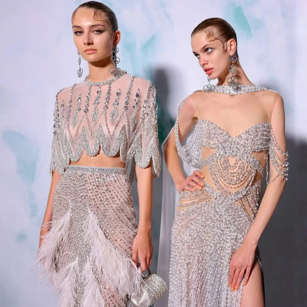 The models wear elaborate silver nude two-piece evening dresses featuring silver beaded details, sheer mesh, and feather accents.