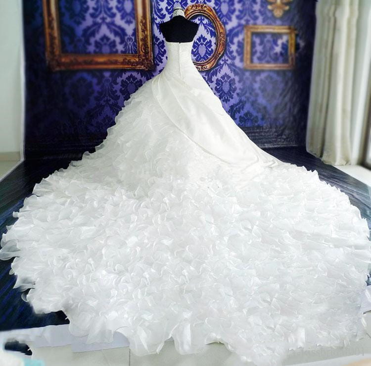 A voluminous white wedding dress wholesale featuring a fitted bodice and an expansive ruffled skirt displayed on a black mannequin against a blue damask backdrop.