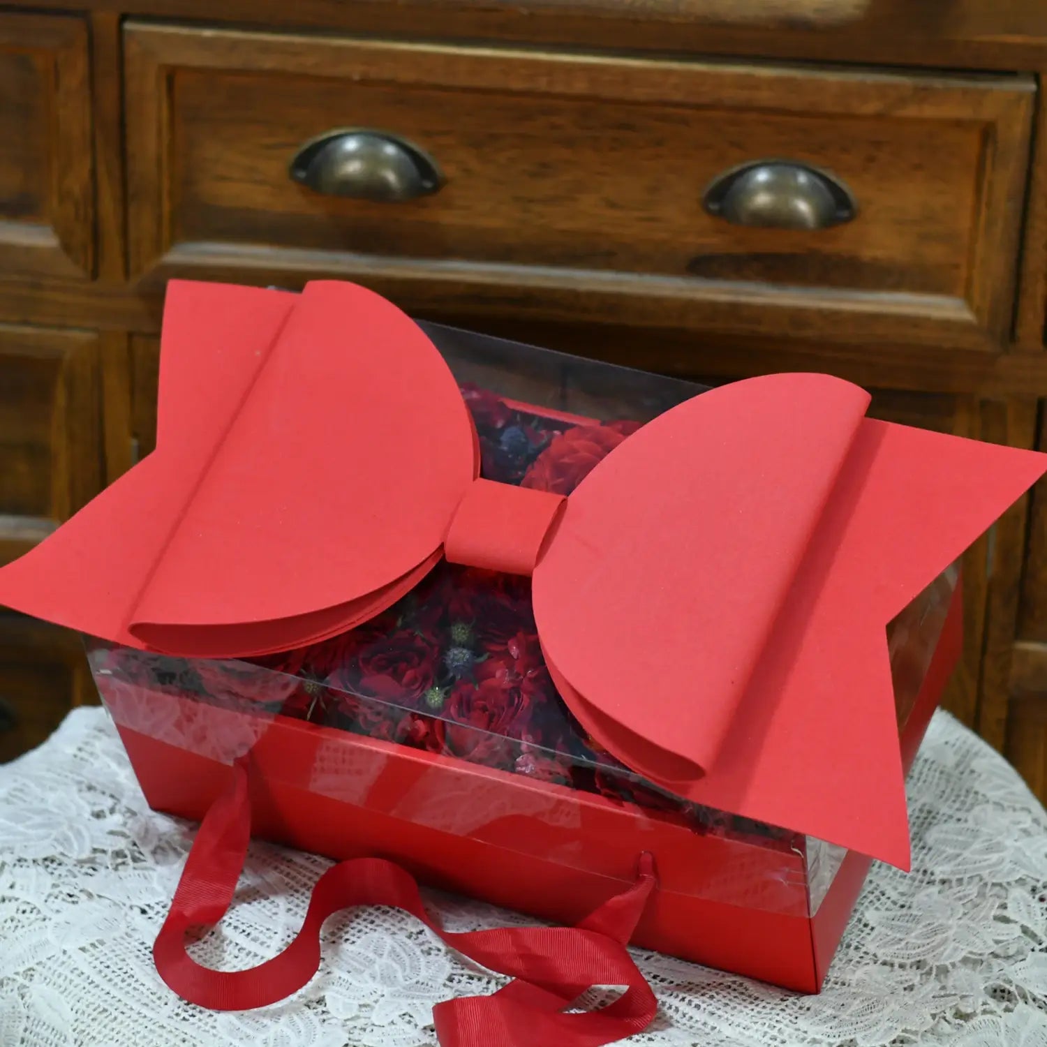 A vibrant red bow knot handheld flower box with a clear window contains a transparent gift box filled with roses.