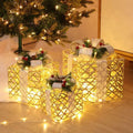 Three golden lattice led gift boxes with warm white led lights white ribbons and festive greenery accents