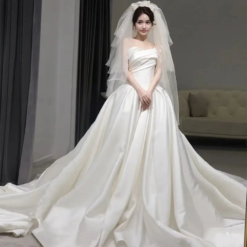 A flowing white temperament princess style wedding gown made of french satin with a strapless bodice, an elaborate train, and a matching sheer veil with delicate floral embellishments.