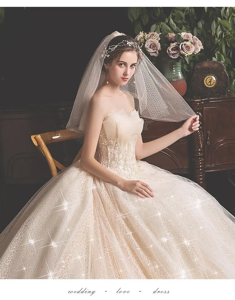 A bride wears an ivory lace summer tube top wedding gown with a fitted bodice and mori style trailing tulle skirt complemented by a delicate veil and sparkling headpiece.