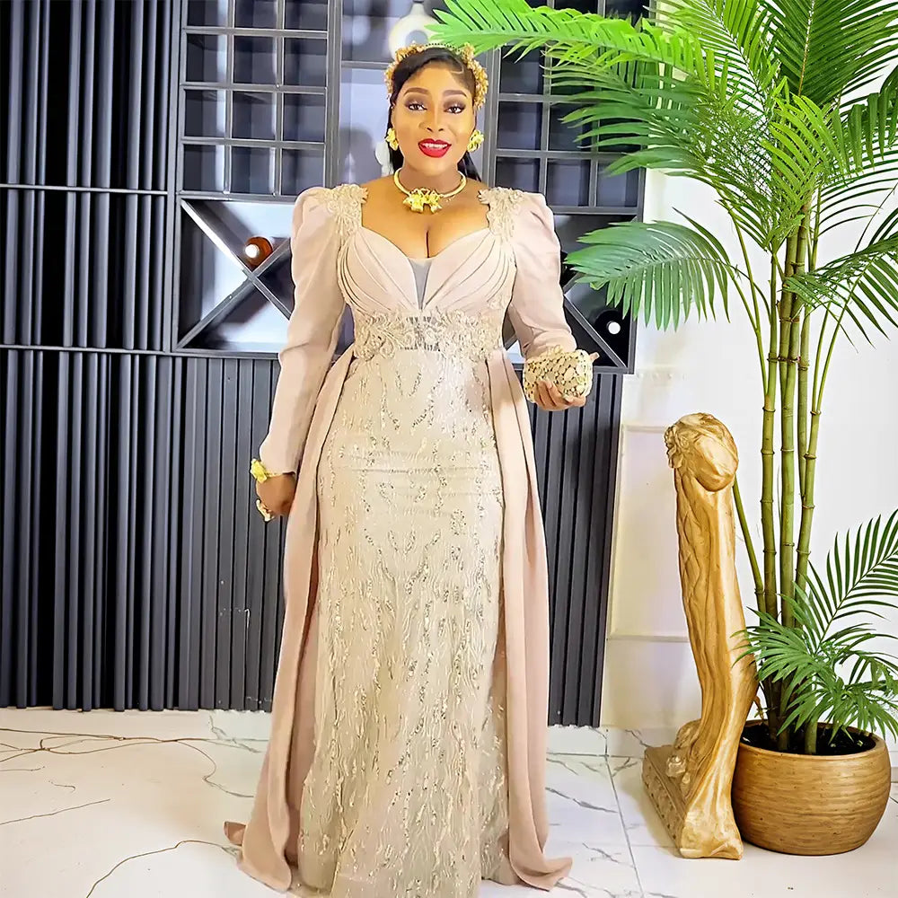 A woman wearing a plus size long sleeve sequined evening dress featuring a light beige lace bodice with long sleeves and a soft blush pink satin skirt.