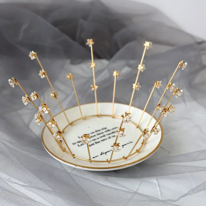 A delicate gold crown with tiny crystal flowers and star-shaped embellishments resting on a white ceramic plate showcasing bride wedding jewelry handmade star design.