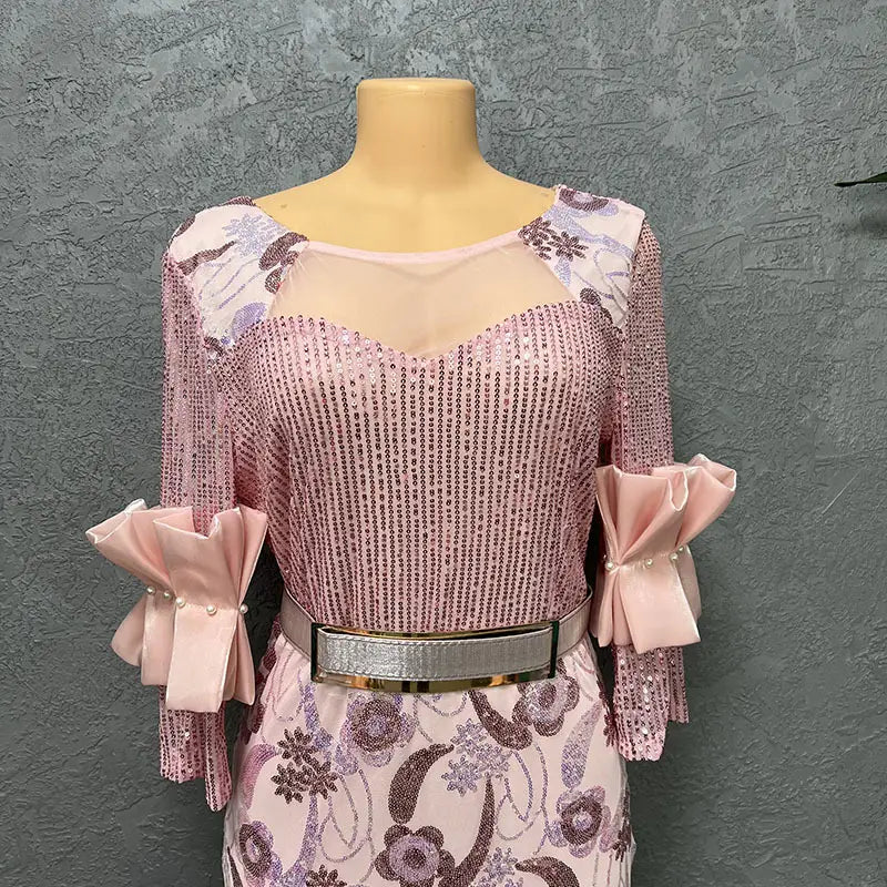 A pink sequined half sleeve slim fit round neck dress with floral patterned sleeves bow cuffs and a silver belt for evening wear.