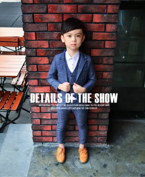A young boy wearing a navy blue plaid children’s three-piece fashion suit with matching trousers, a white collared shirt, and tan leather shoes showcasing a stylish comfort suit.