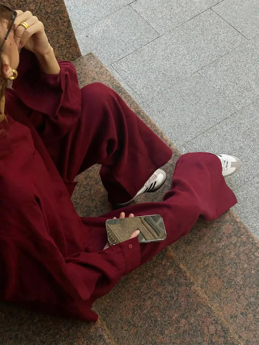 a person wearing a deep red long sleeve cotton shirt trousers suit seated on stone steps displaying a loose-fitting shirt and wide-leg trousers