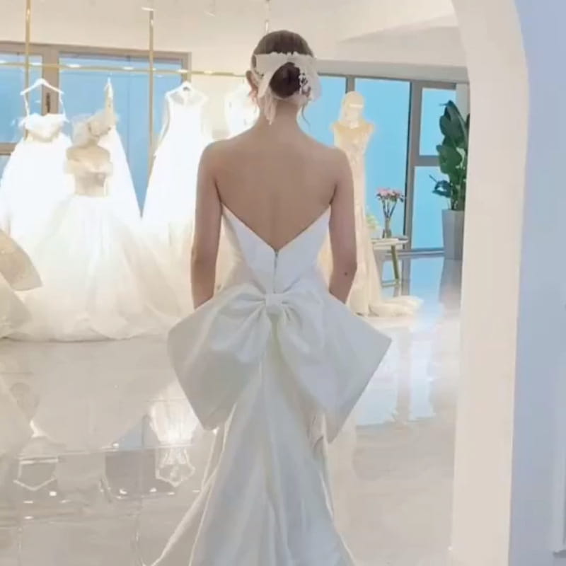 A woman wears the back side of a white satin small trailing temperament mermaid wedding dress featuring an open back and a large bow at the waist.