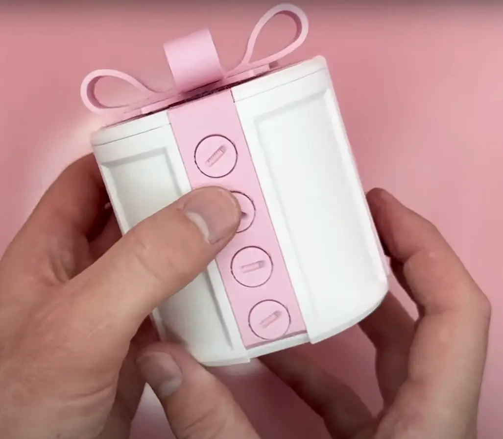 A white cube-shaped gift box with a pink ribbon bow and a vertical strip of pink paper featuring circular button designs, ideal as a creative day gift box for valentine day.