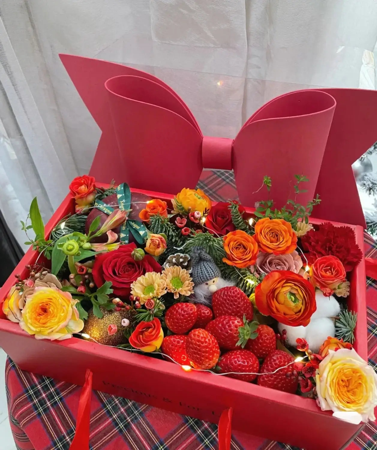 a vibrant red bow knot handheld flower box with a clear window featuring a large glossy paper bow and filled with fresh orange yellow and deep red flowers nestled among strawberries greenery and festive ornaments