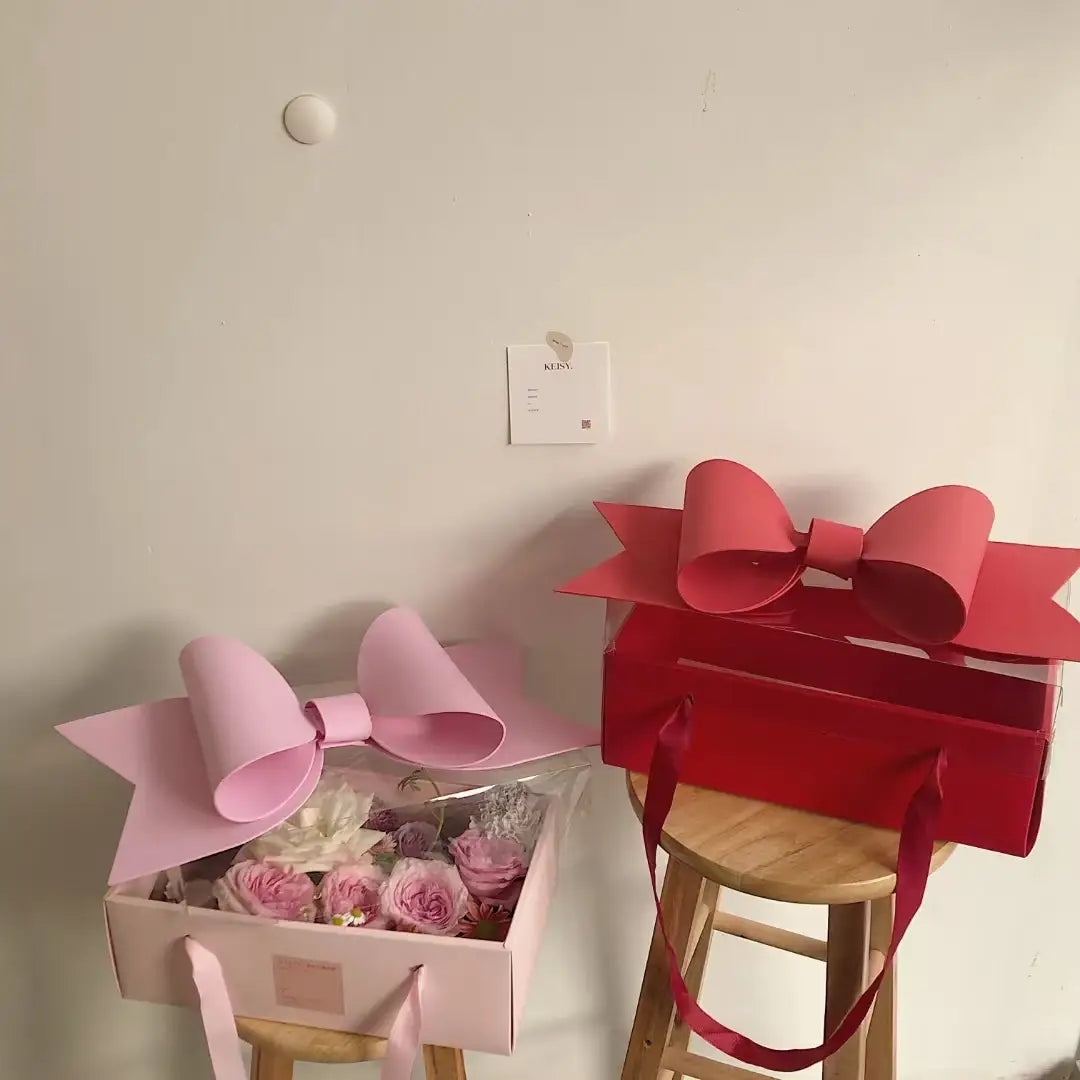 A pink bow knot handheld adorns a red handheld flower box filled with roses and featuring a clear window.