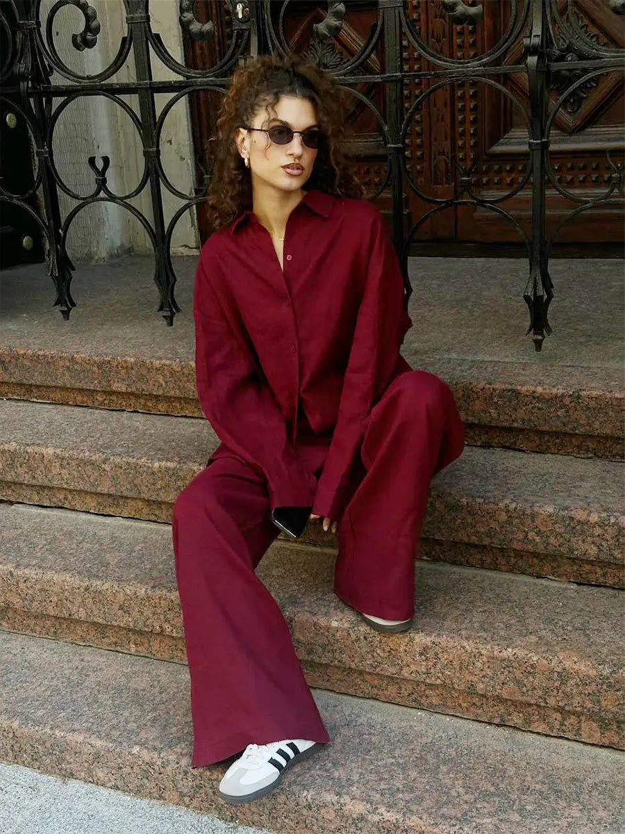 a woman wearing a burgundy long sleeve cotton shirt trousers suit with an oversized button-up shirt and matching wide-leg pants paired with white adidas sneakers