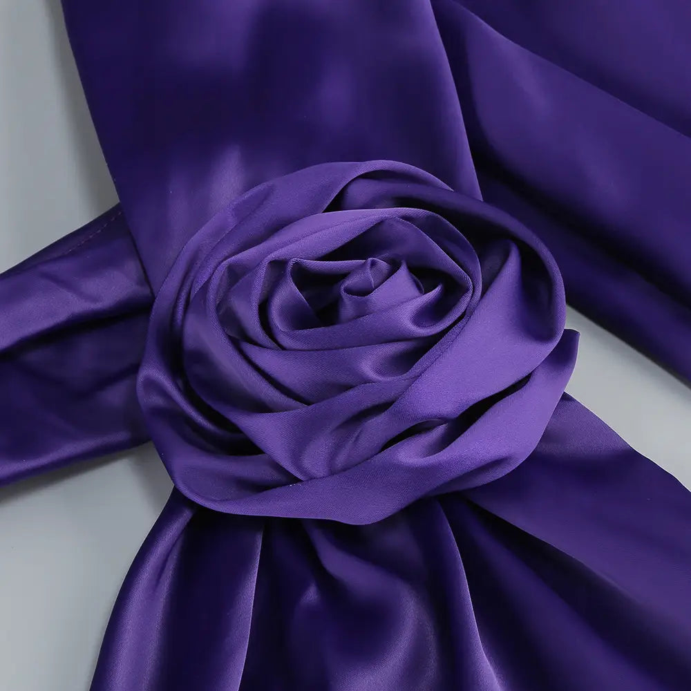 A luxurious deep purple satin scarf with a rose-shaped embellishment elegantly draped on a one shoulder fitted high waist purple satin rose gown highlighting the waist pure color.
