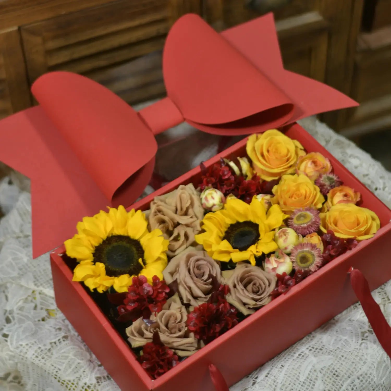 red rectangular handheld flower box with clear window featuring a large bow knot and filled with preserved yellow sunflowers beige roses and red flowers