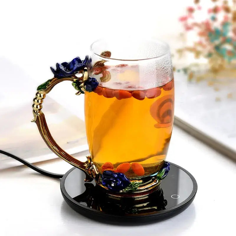 a clear glass mug cup warmer with an ornate gold-toned handle and base featuring blue floral embellishments, filled with amber tea garnished with red goji berries.