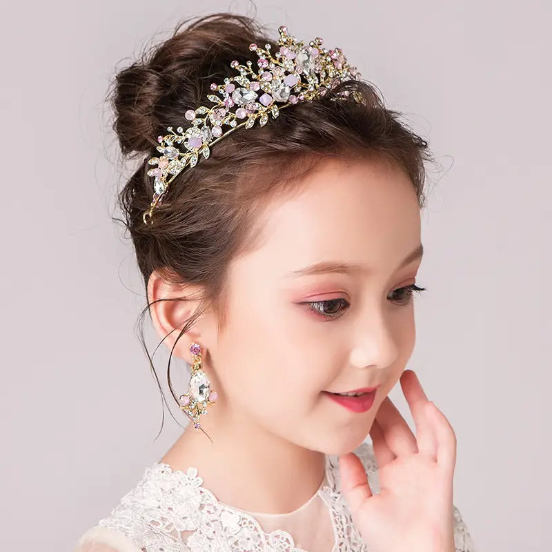 A delicate gold-toned princess crown tiara adorned with sparkling clear and soft pink crystals features an intricate floral design for a catwalk cute princess look.