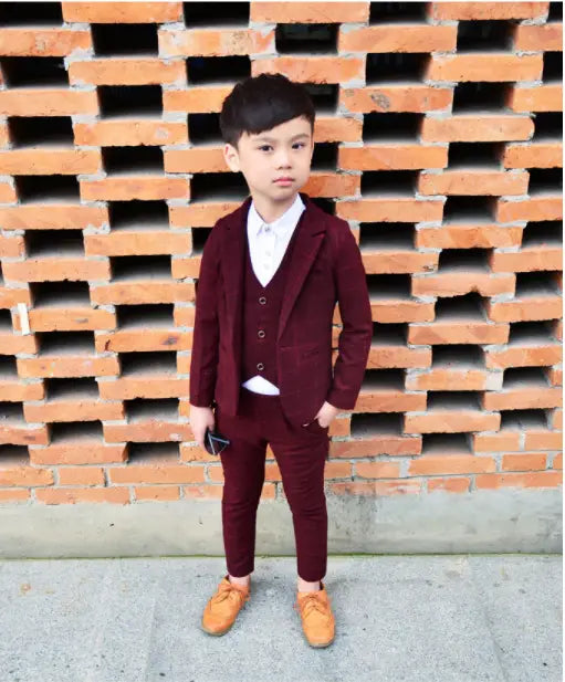 A young boy models a maroon children’s three-piece fashion suit with matching trousers and vest paired with tan shoes showcasing a comfort suit alternative to the blue plaid children’s three-piece fashion suit.