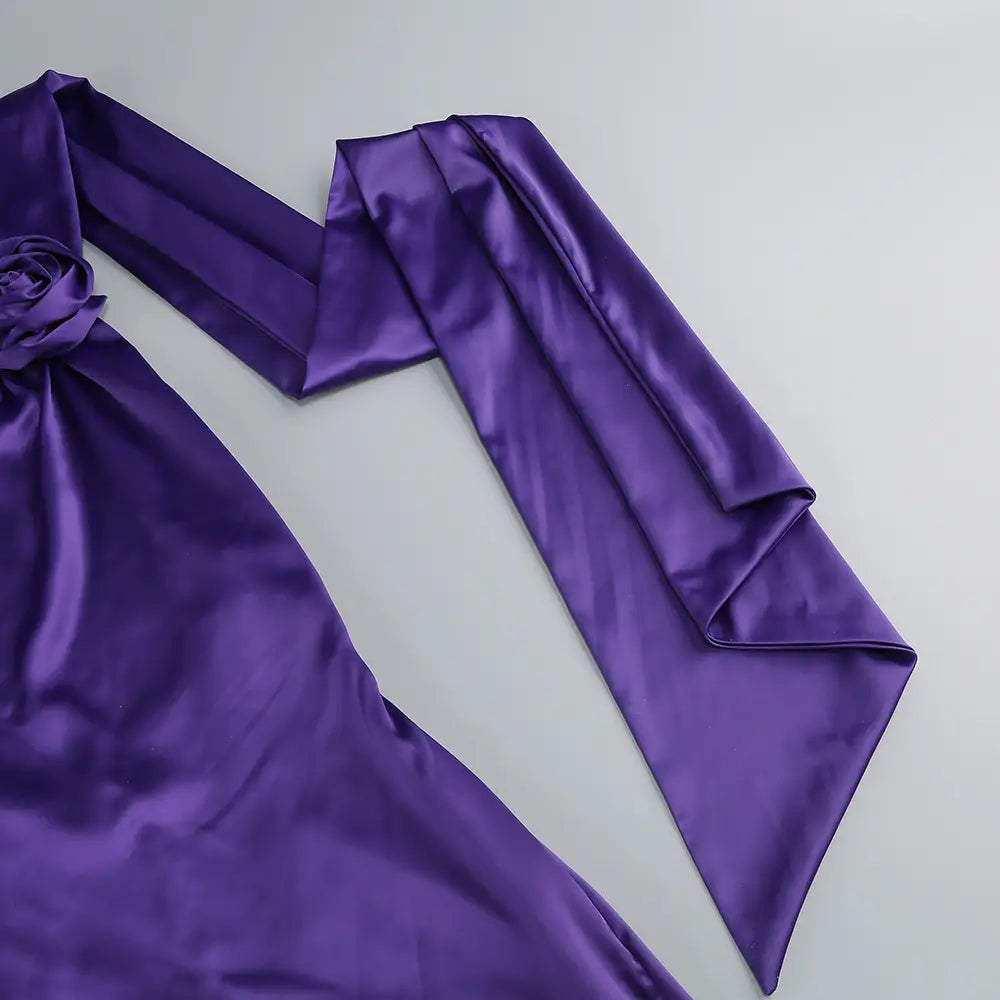 A rich purple satin one shoulder fitted high waist gown with a flowing elegant design and a decorative rose accent at the neckline.