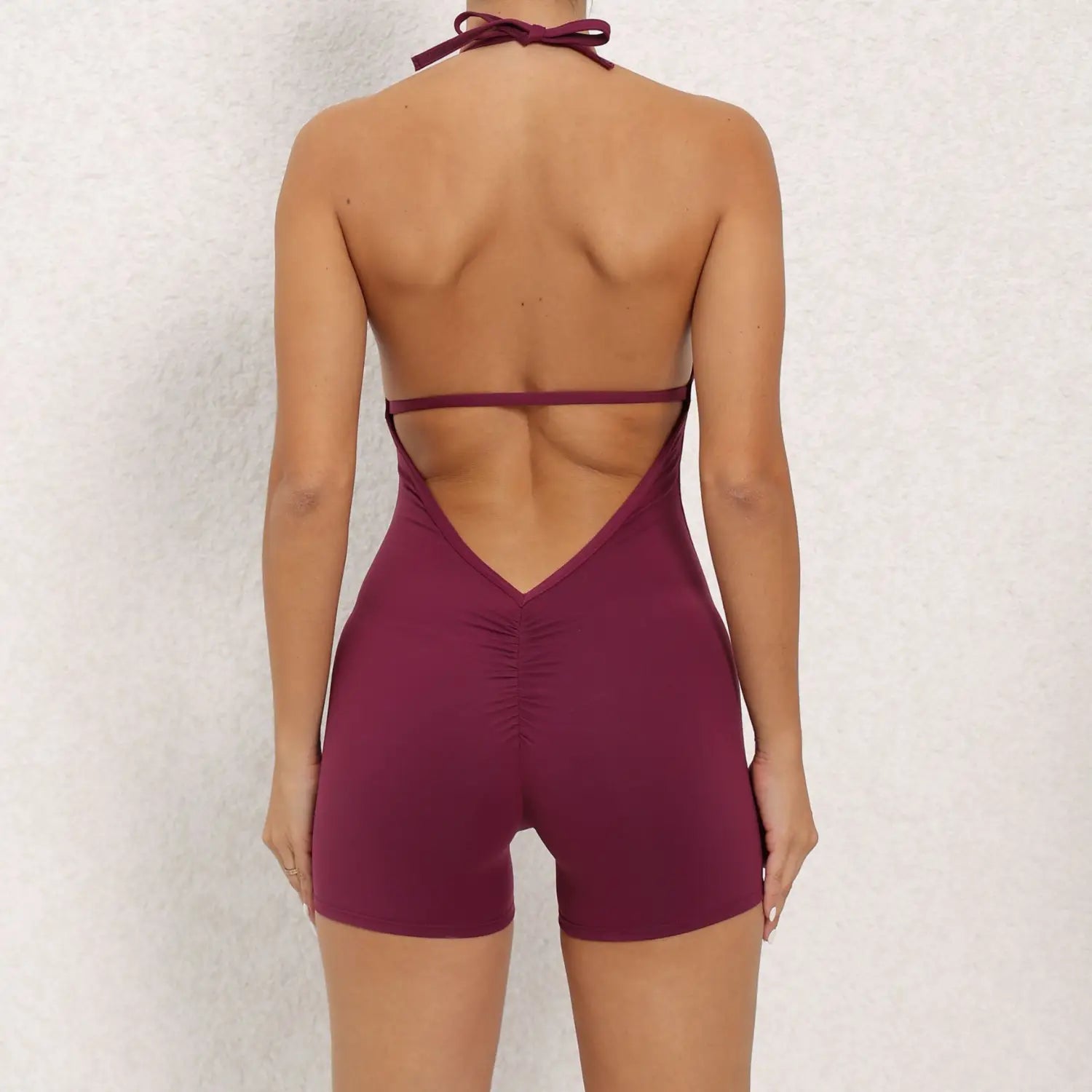 a maroon halter neck high elastic one-piece yoga romper featuring a deep back cutout and ruched waistband shown from the back side
