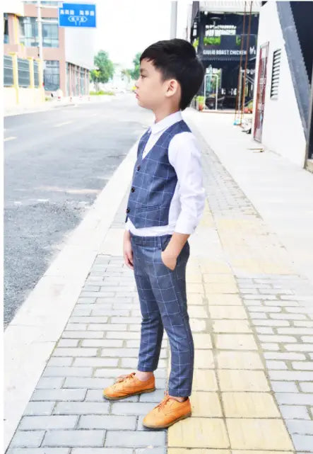 A young boy wearing a navy blue plaid children’s three-piece fashion suit with a comfort suit vest, white shirt, matching trousers, and tan leather shoes.