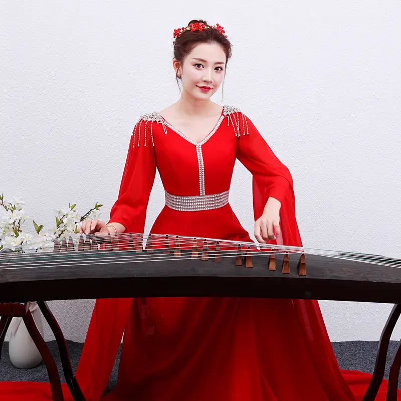 A woman wearing a vibrant red long guzheng performance dress with silver beading and long flowing sleeves sits gracefully beside an elegant black guqin.