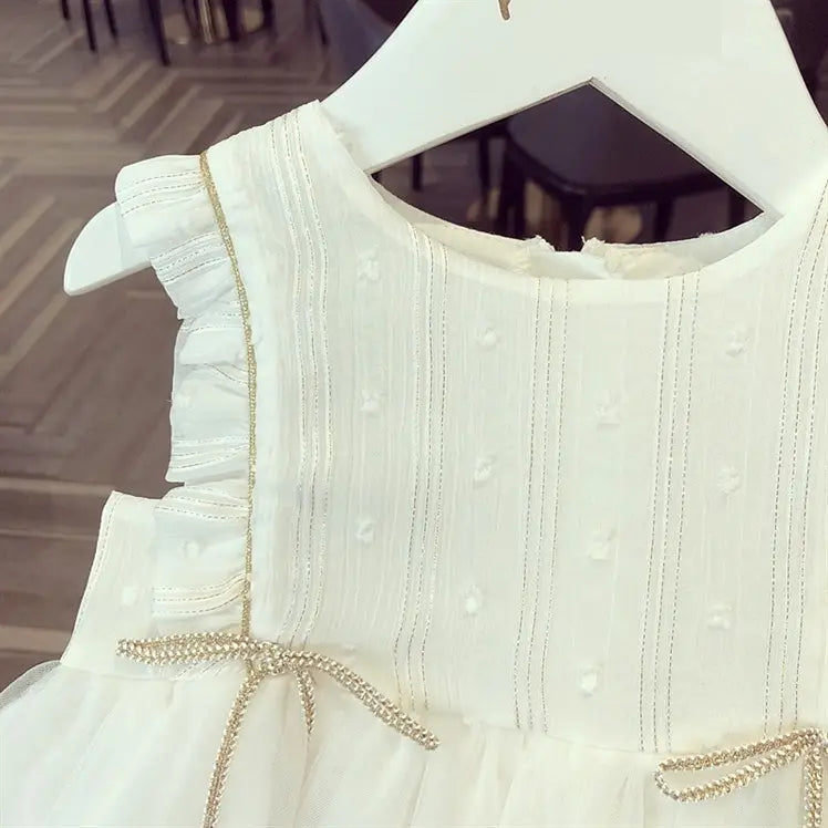 White sleeveless baby gauze princess dress with delicate ruffled shoulders, subtle gold trim, and bow at the waist ideal as a gauze dress summer outfit.