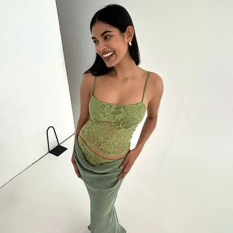 A woman wears a sage green lace backless tube crop top with thin spaghetti straps and a matching satin sling stitching ruffle skirt featuring stitching ruffle hip details.
