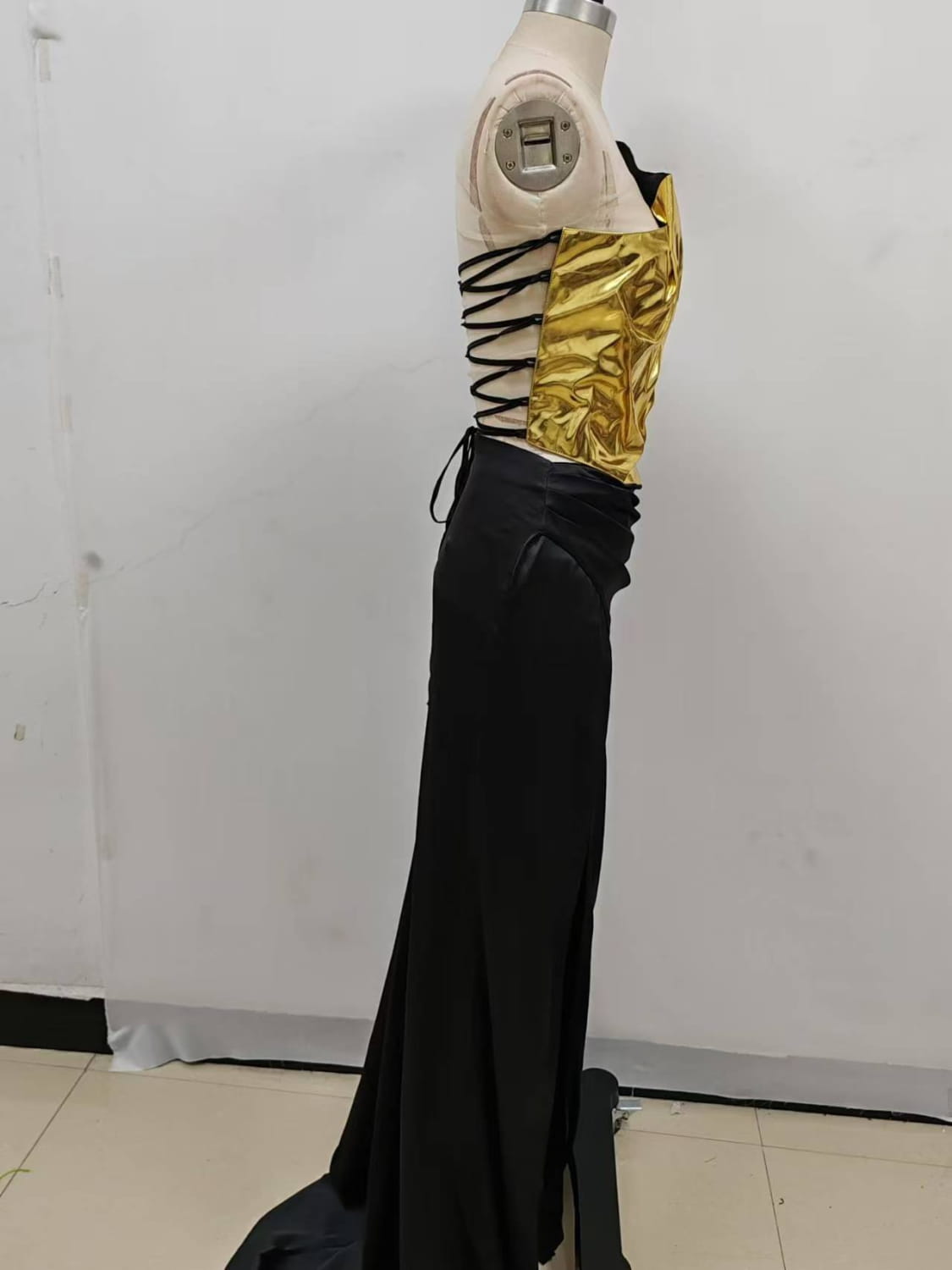 A striking one-shoulder slit mop black two-piece set featuring a black and gold corset top with glossy metallic finish paired with high-waisted black satin trousers showcasing set sexy lacing details.