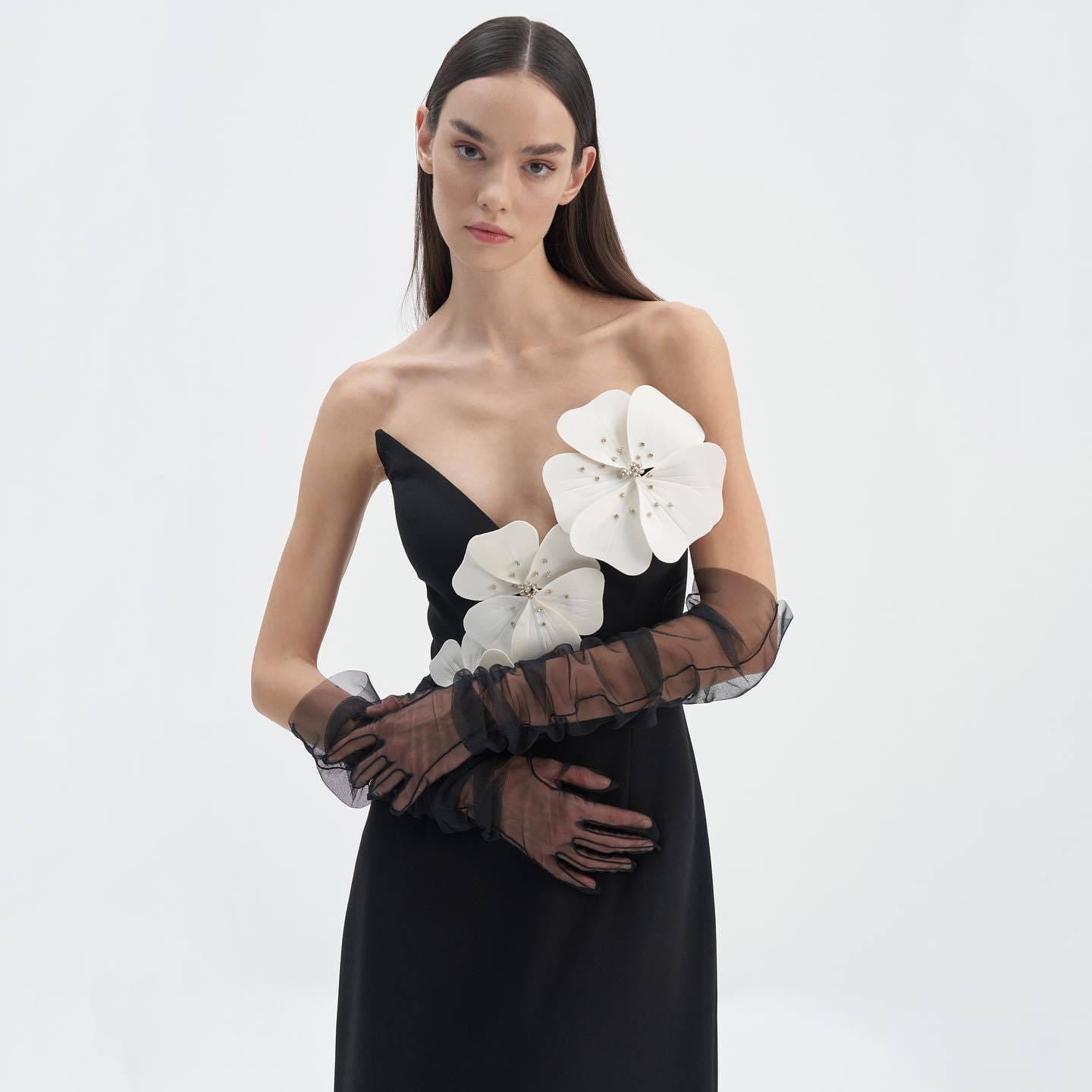 black backless bandage gown featuring a black strapless design adorned with large white floral appliqués and sheer brown lace gloves showcasing the bandage chest white detail