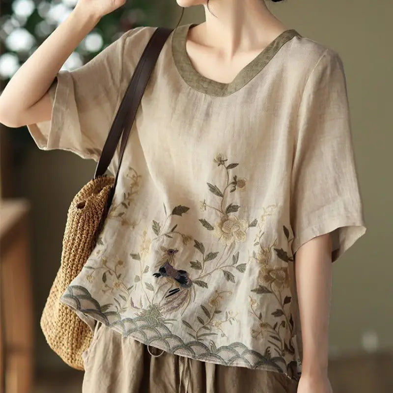Beige linen round neck top with delicate art retro embroidery of floral and birds in earthy tones, featuring a contrasting olive green colored round neck collar and hem.