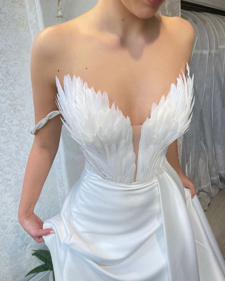 A white satin wedding gown with a plunging neckline and angel wing embellishments featuring feathers sweeping the floor, exemplifying luxury women’s fashion.