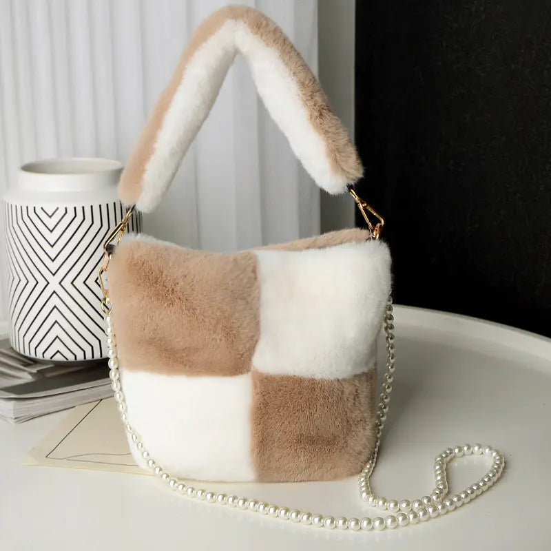 A cozy beige and white checkerboard plush bucket bag made of soft faux fur featuring a matching fluffy handle and an elegant pearl chain design.