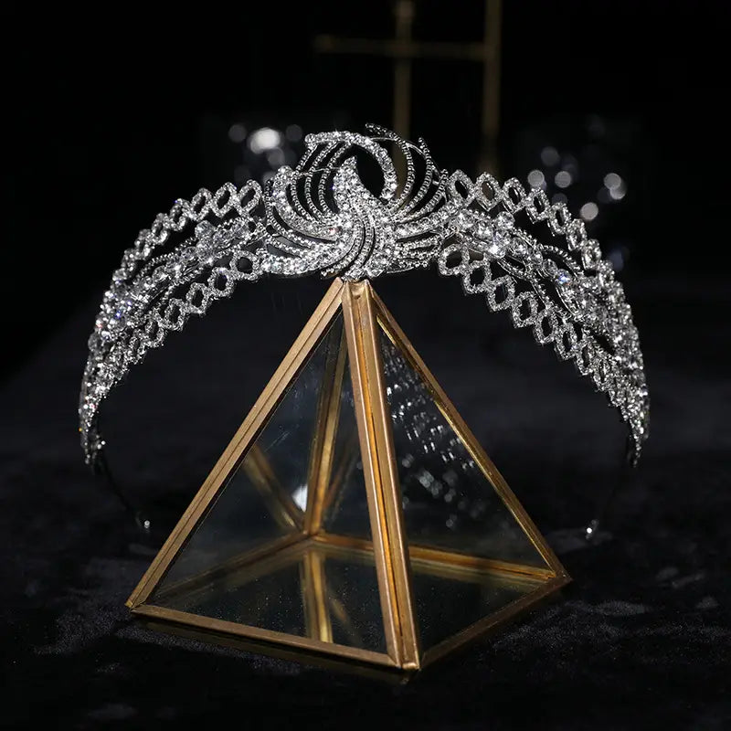 a sparkling silver rhinestone crown luxury bridal headdress with intricate diamond-like crystals and a central phoenix motif elegantly displayed on a gold-framed glass pyramid stand