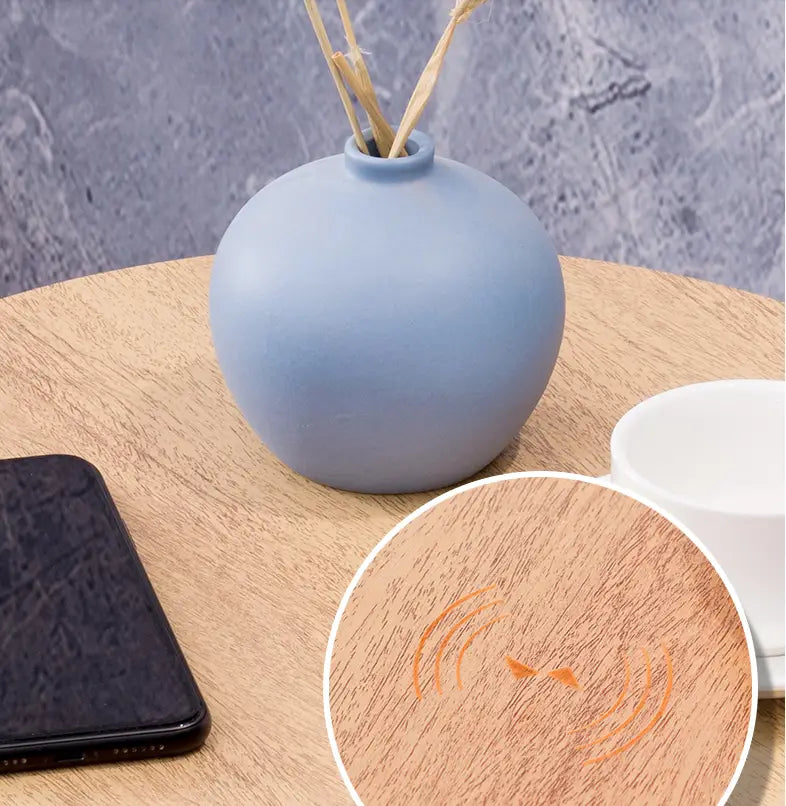 a matte light blue ceramic diffuser vase with a rounded body and narrow neck holding dried reed sticks on a log color smart side table with wireless charging and smart voice features