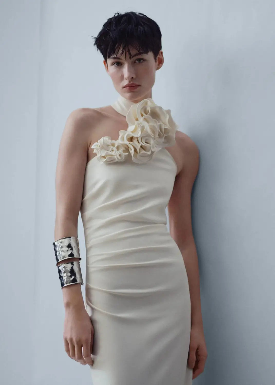 a sleek off-white satin one-shoulder gown featuring a ruched bodice and an elaborate white ribbon flower halter embellishment at the neckline