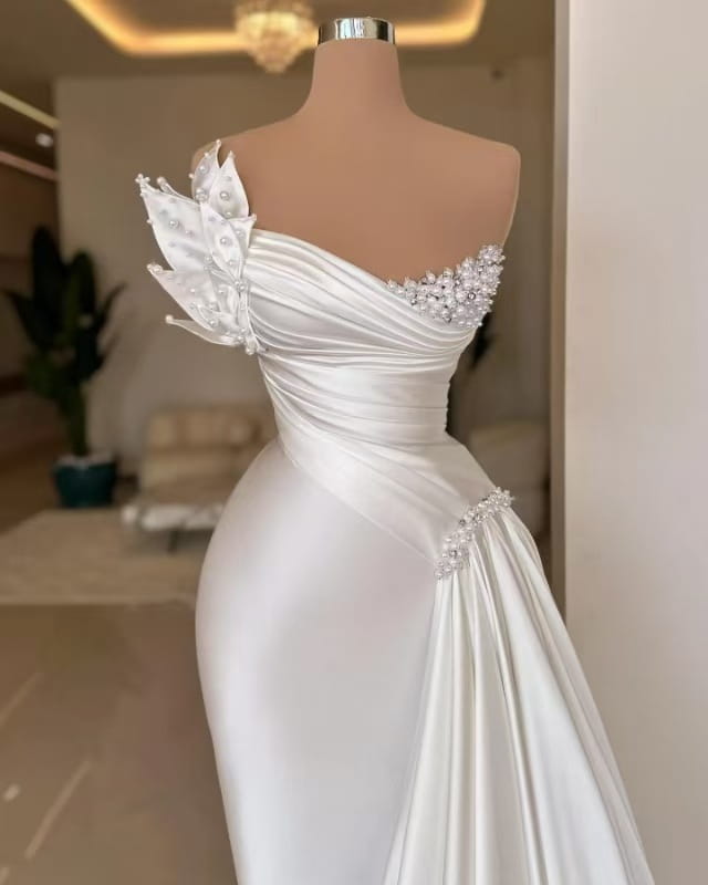 A white graceful satin off-the-shoulder wedding dress featuring a sleek one-shoulder design with sparkling beaded embellishments and delicate pearl accents on the shoulder and waist.