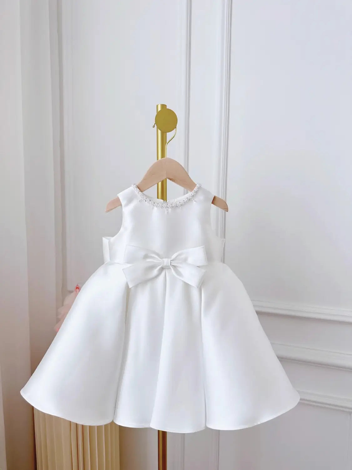 A white satin sleeveless baby dress white classic style featuring a beaded neckline and a large bow at the waist.