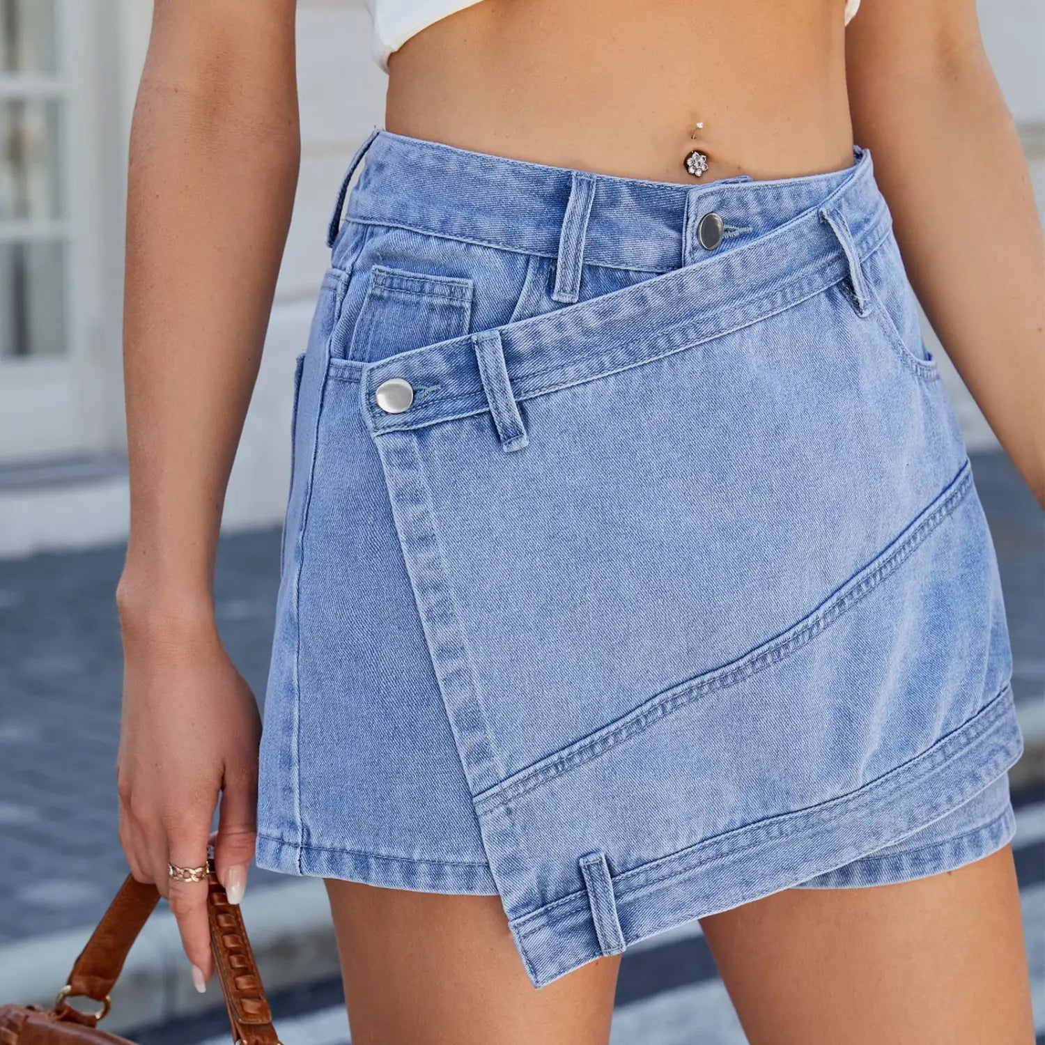 Light-wash denim short skirt with high waist, diagonal flap pocket, silver-toned button closures, and washed irregular denim detailing.