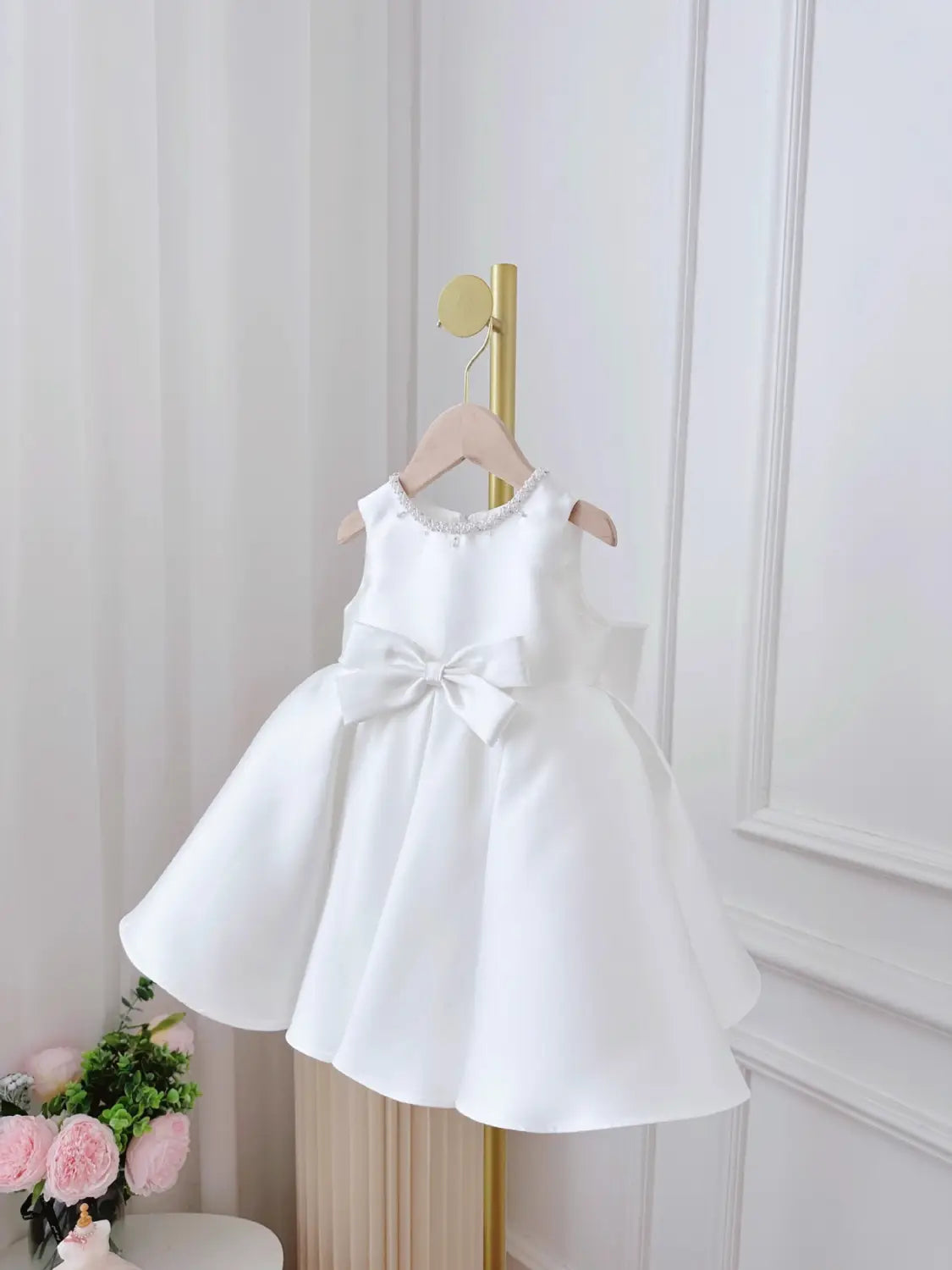 A white satin sleeveless baby dress white classic style featuring a beaded neckline and a large bow at the waist.