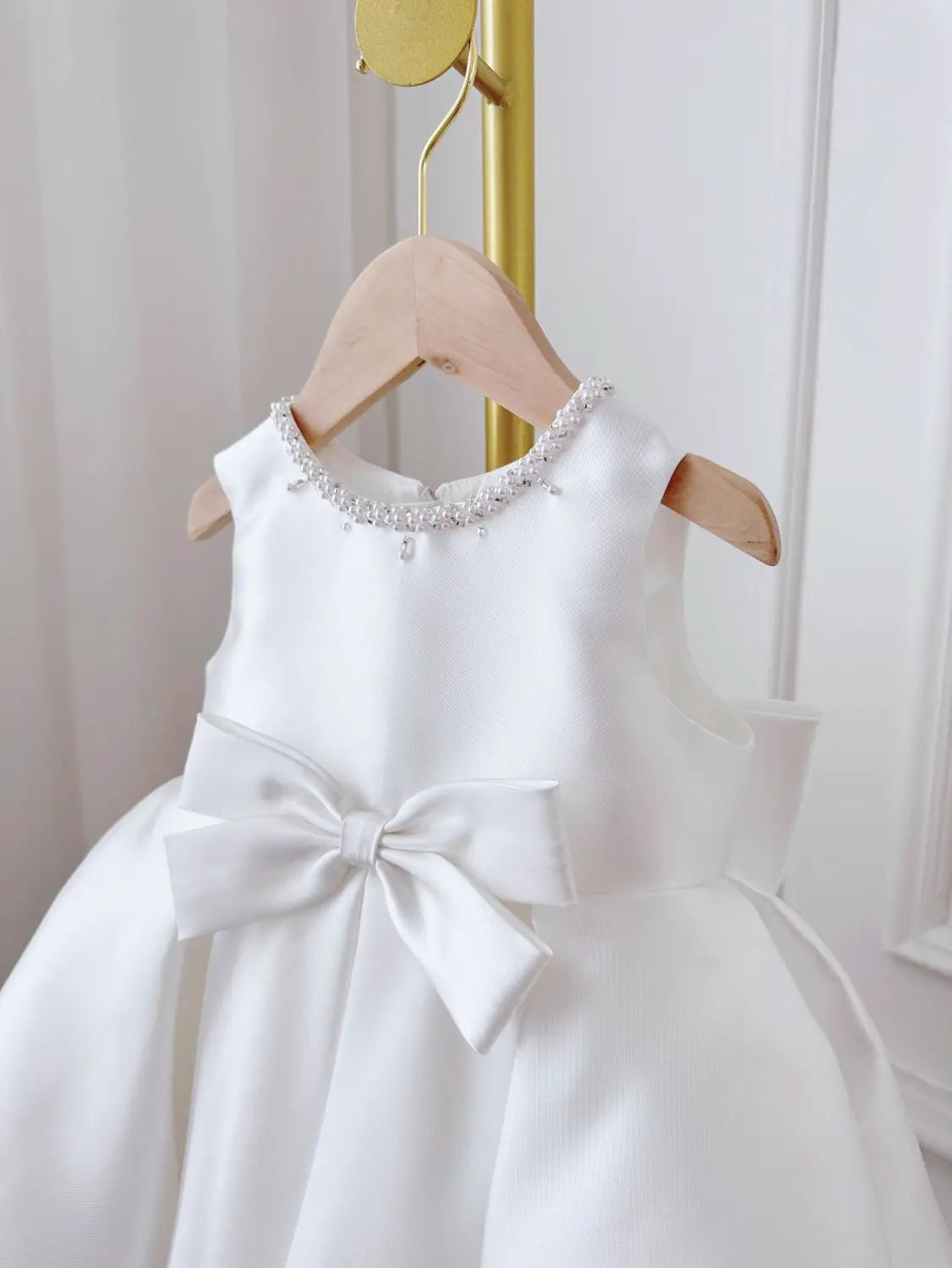 A white satin baby dress white with long sleeves featuring a beaded neckline and a large bow at the waist in a white classic style.