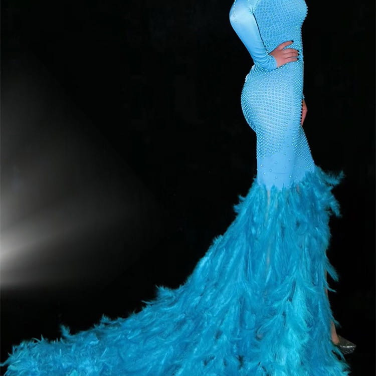 A striking turquoise mermaid-style gown with a fitted bodice and long sleeves adorned with intricate beading and a voluminous water bottle feather trailing train.