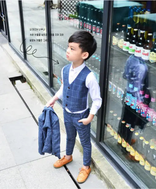 a young boy wearing a navy blue plaid children’s three-piece fashion suit consisting of a vest, white collared shirt, matching trousers, and tan leather shoes for a stylish comfort suit look