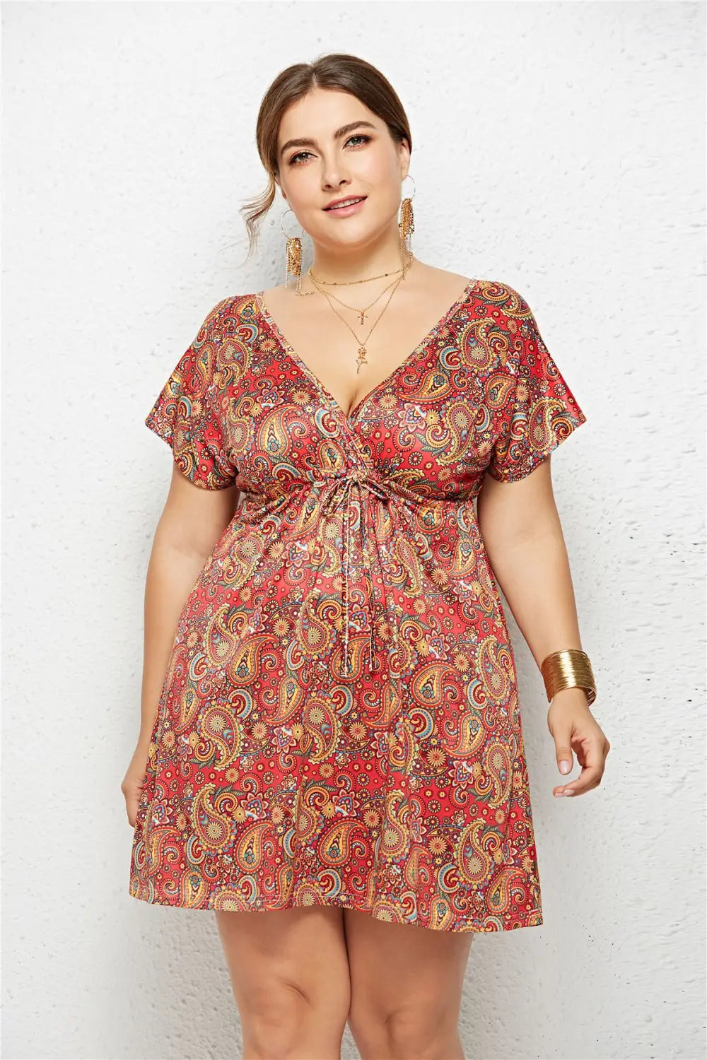 A plus-size woman wears a vibrant red geometric printed deep v-neck dress with an elastic waist and short flutter sleeves featuring an intricate paisley pattern in gold and green.