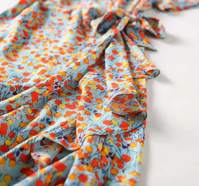 A flowing light blue french floral slim polka dot wrap dress adorned with a vibrant pattern of orange yellow and purple abstract flowers.