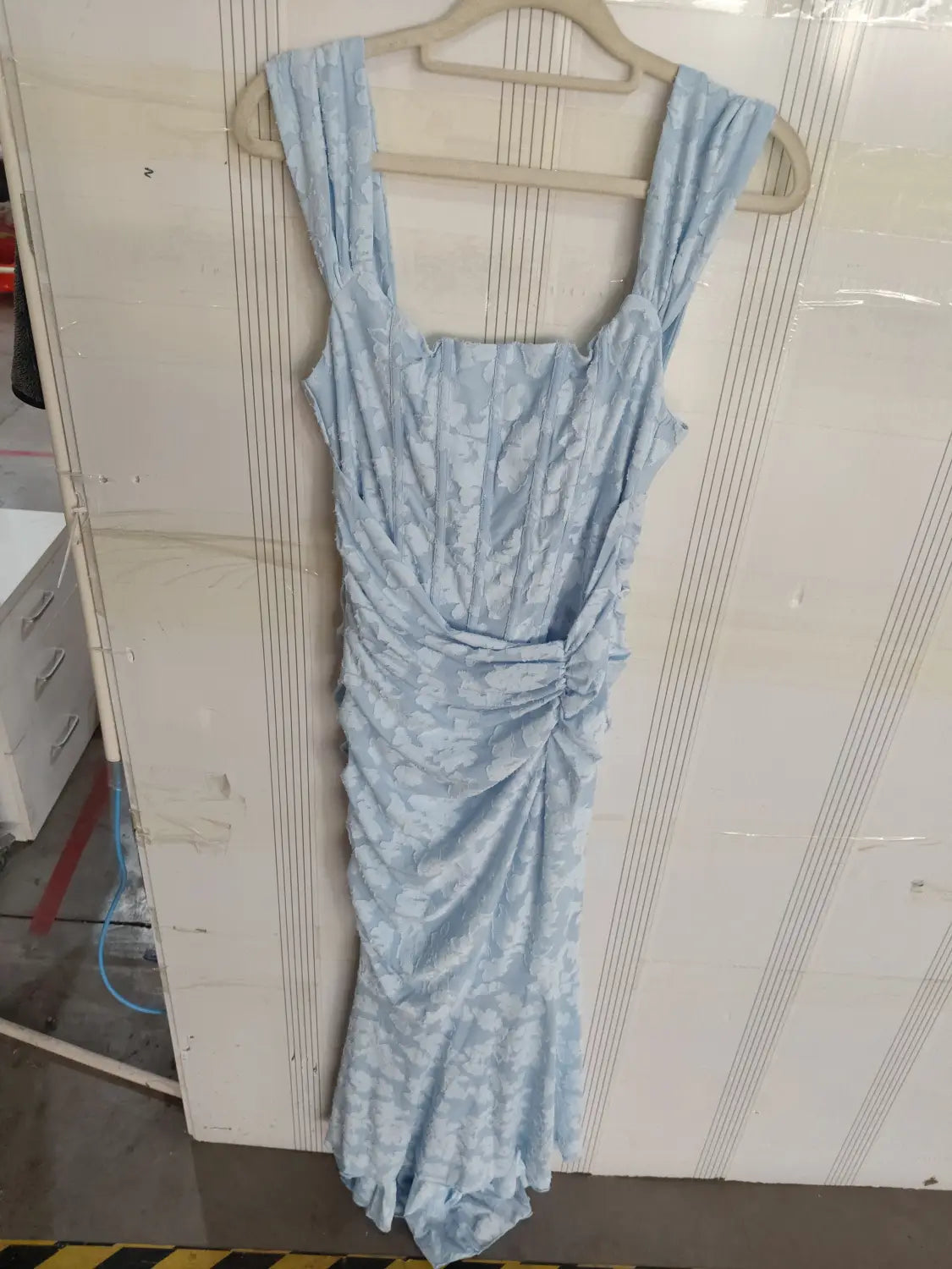 a light blue sleeveless dress with a subtle floral pattern and ruched detailing at the waist displayed on a wooden hanger, ideal as a backless sexy evening dress or women purple sleeveless option.