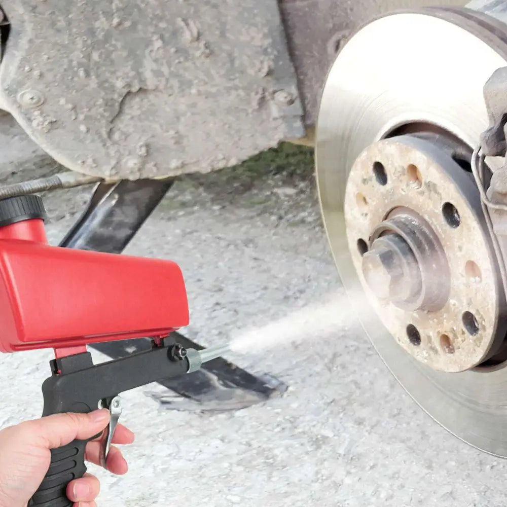 Red and black handheld air portable sandblasting gun applying white paint to a car brake disc for precise coating