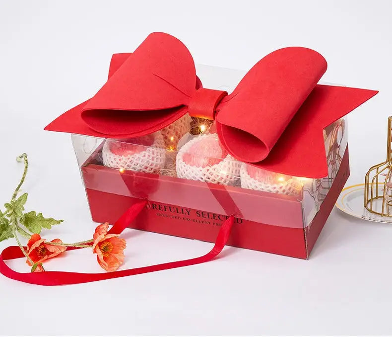 A red bow knot handheld flower box with clear window features a large plush velvet bow on top and clear sides revealing pink fruits wrapped in white netting inside.
