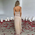 A woman wears a beige one-shoulder backless slit satin gown with thin diagonal straps and an open back view