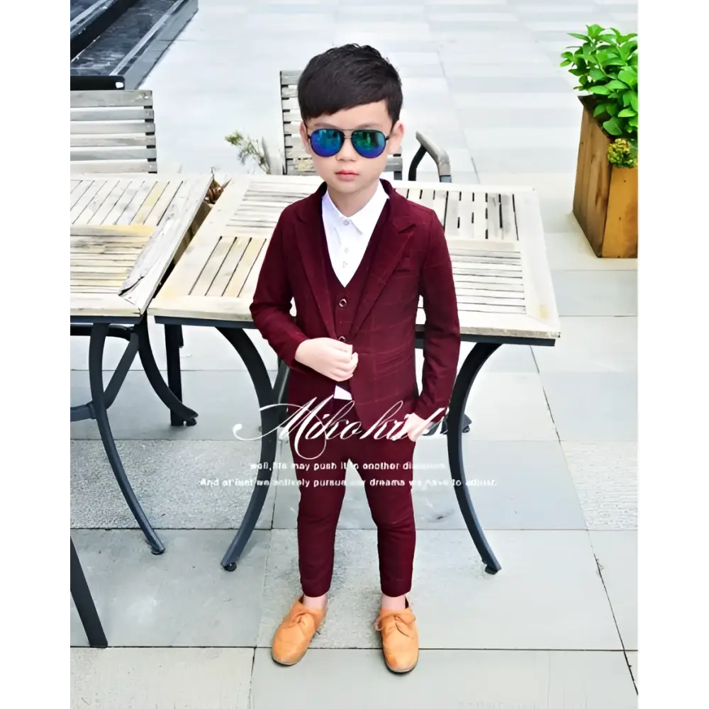A young boy wearing a maroon velvet children’s three-piece fashion suit with matching trousers and a white shirt