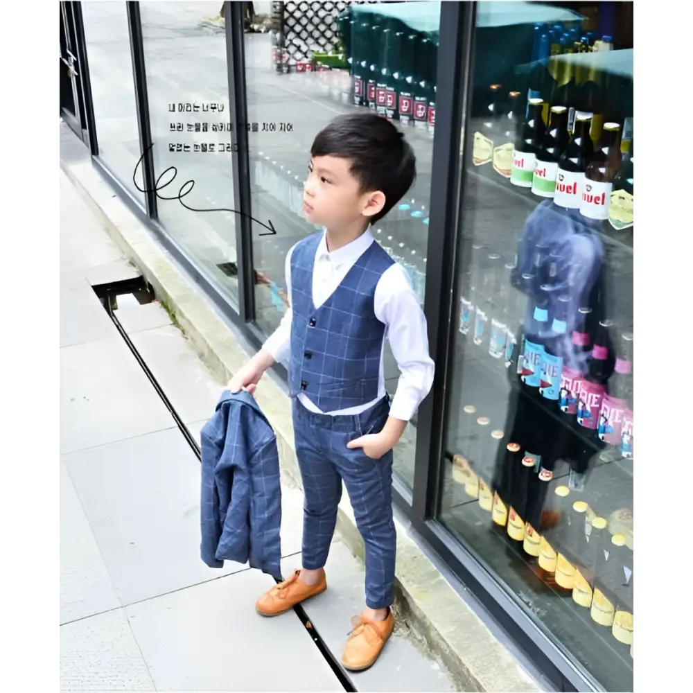 A young boy wearing a blue plaid children’s three-piece fashion comfort suit with matching trousers and orange shoes